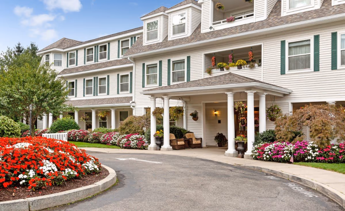 Front entrance of a white multi-story residential building with a covered portico, outdoor seating, and colorful flower beds.