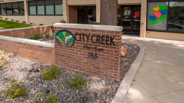 Exterior view of City Creek Post Acute facility showing a brick sign with the facility's name and address number 165. The building entrance is visible in the background with glass doors and windows, and colorful heart-shaped decorations are displayed on one window.