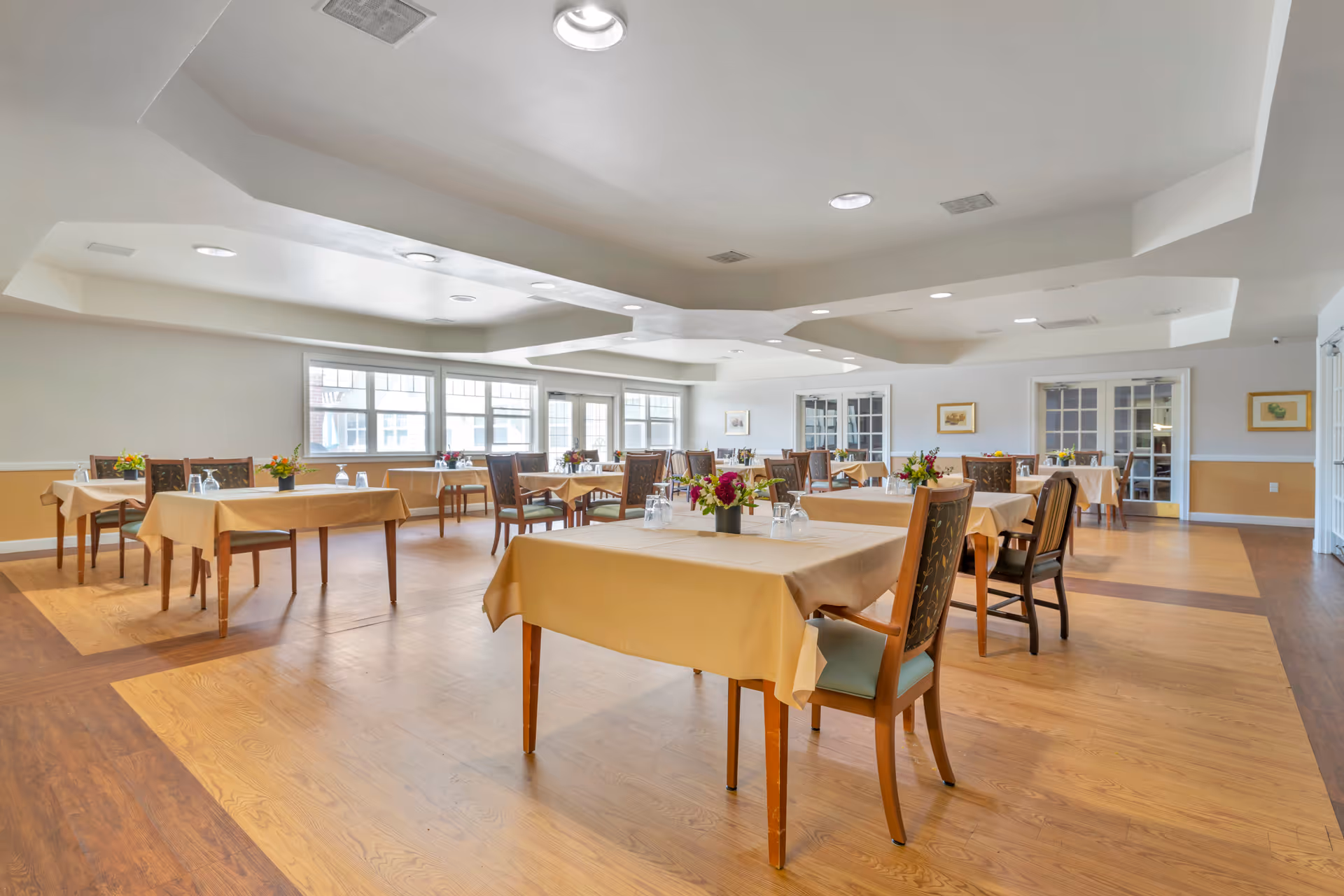 A spacious dining room with multiple tables covered in beige tablecloths, each set with glasses and small flower arrangements. The room has large windows allowing natural light to fill the space, light-colored walls, and a decorative ceiling with recessed lighting. Wooden chairs with cushioned seats surround the tables.