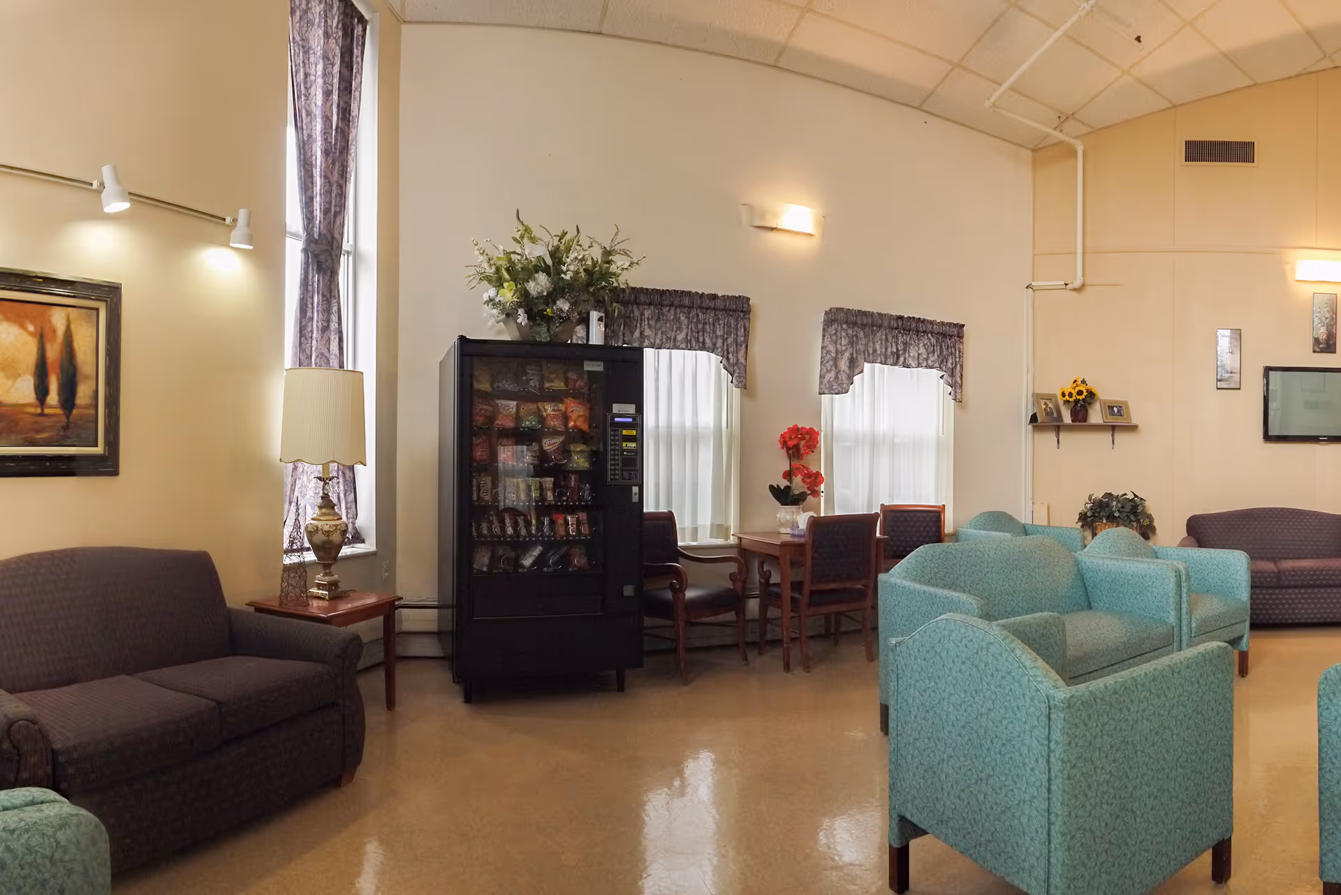 A common area in a senior living facility featuring multiple upholstered chairs and sofas arranged around the room. There is a vending machine against the wall, a small table with chairs near windows with curtains, a table lamp on a side table, and decorative flowers and framed artwork on the walls.