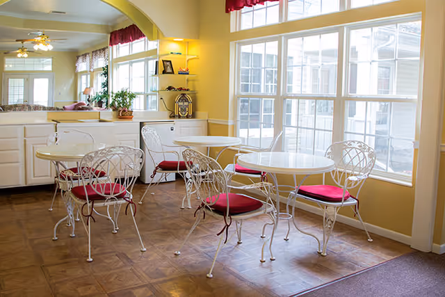 Bright seating area with white metal bistro tables and chairs with red cushions beside large windows in a senior living facility.