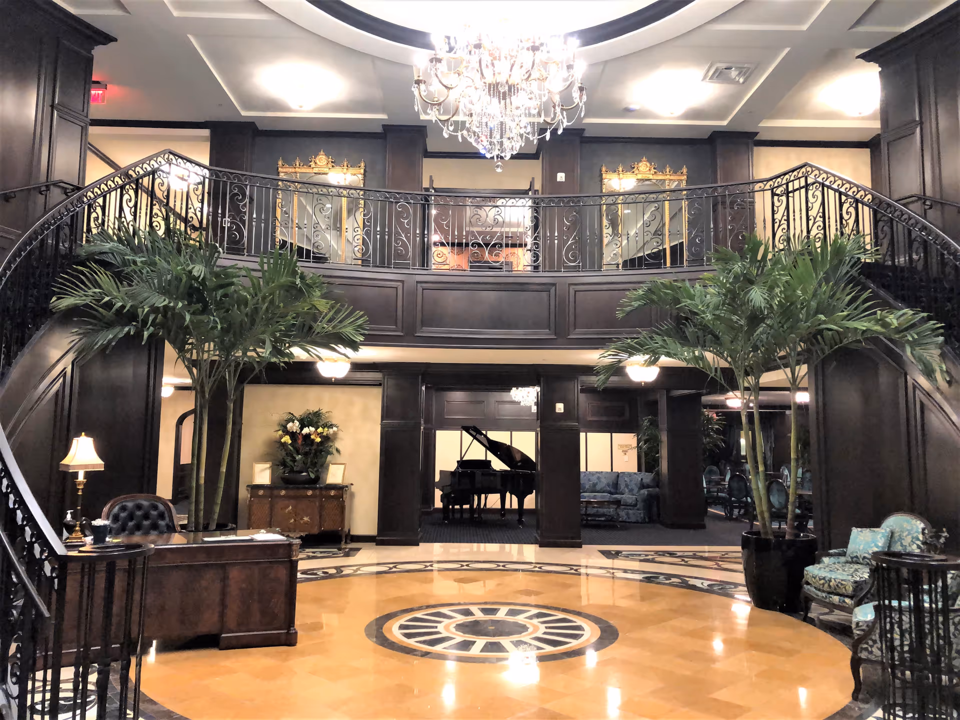 Elegant interior lobby area with a grand chandelier hanging from the ceiling, a curved staircase with ornate railings, large potted plants, a wooden reception desk with a lamp, and a grand piano in the background. The space features dark wood paneling, decorative mirrors, and comfortable seating areas.