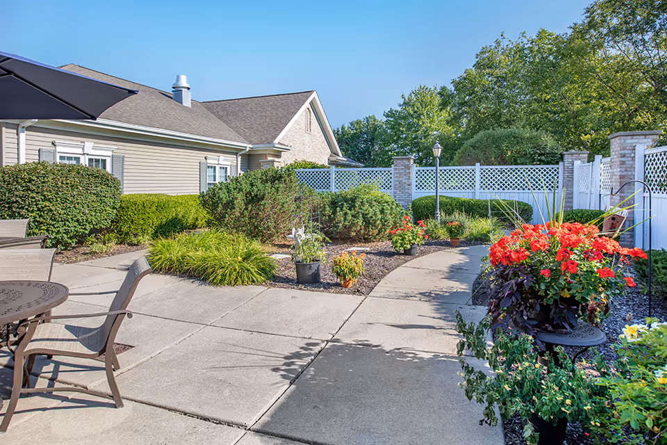 Sunny outdoor courtyard with a paved walkway, patio seating, colorful flowering planters, shrubs, and a low building with a white lattice fence.