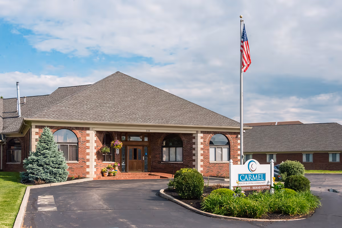 Front entrance of a brick senior living facility with a landscaped circular drive, an American flag, and a sign reading Carmel.