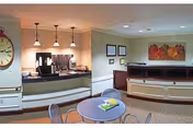 Community lounge with a round table and chairs in the foreground and a coffee/serving counter with pendant lights and artwork on the back wall.