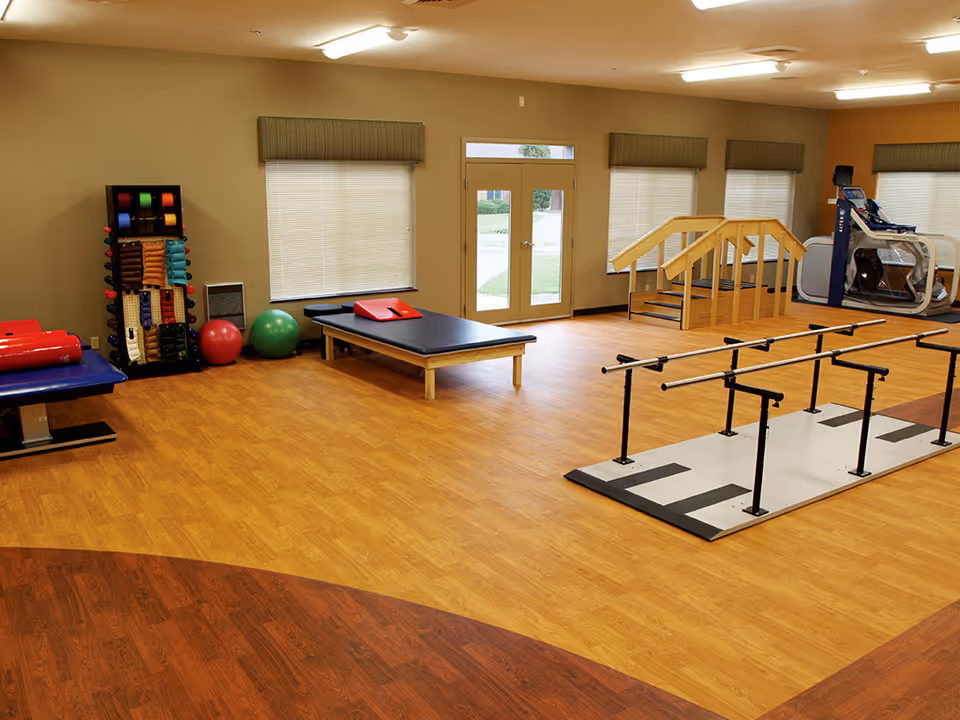 A spacious physical therapy room with wooden flooring, featuring parallel bars for walking exercises, a set of wooden stairs for rehabilitation, exercise balls, a treatment table, and various therapy equipment. Large windows with blinds and a glass door provide natural light.