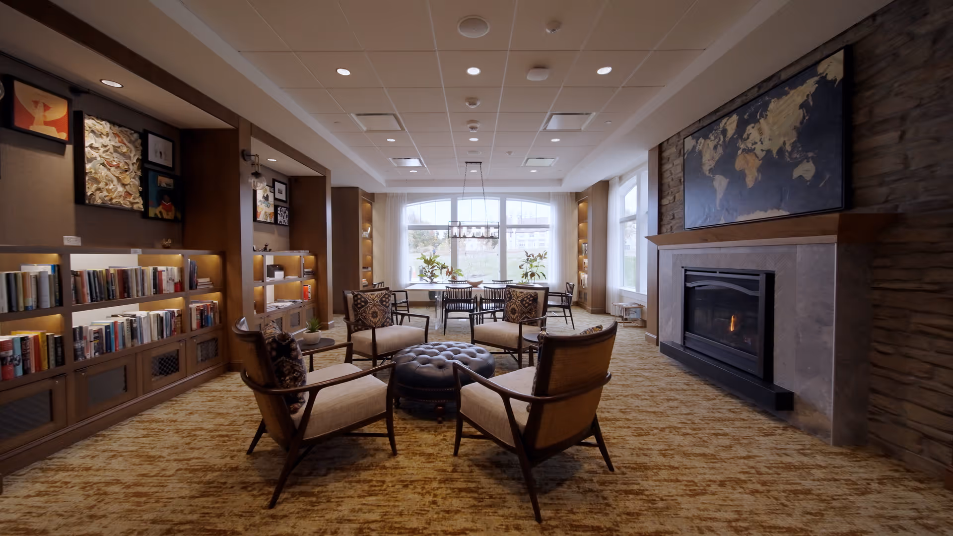 A cozy senior living common area with four cushioned armchairs arranged around a round tufted ottoman. The room features a fireplace with a world map above it on the right wall, built-in bookshelves filled with books and decorative items on the left wall, and a large window at the far end letting in natural light. There is a dining table with chairs near the window and warm, neutral-toned carpeting throughout.