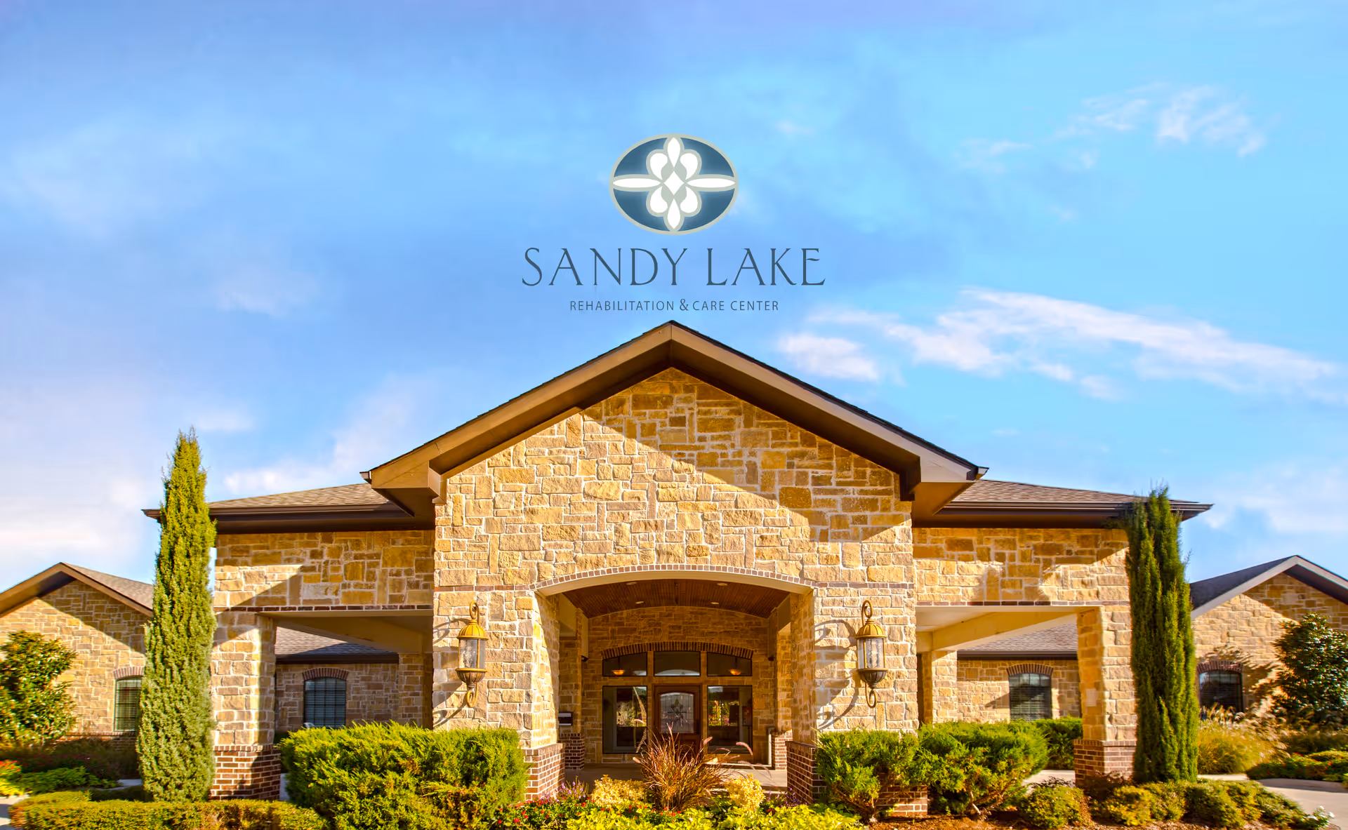 Front exterior view of Sandy Lake Rehabilitation and Care Center, a stone building with a peaked roof, surrounded by greenery and tall shrubs under a blue sky.