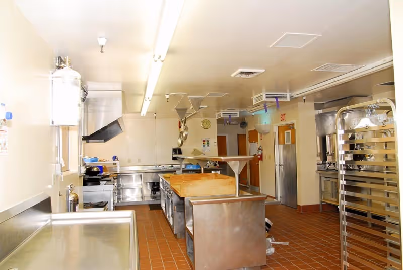 A clean, well-lit commercial kitchen with stainless steel counters, cooking equipment, and a central island with a wooden cutting board. There are overhead ventilation hoods, a fire extinguisher, and a metal rack on wheels. The floor is tiled with reddish-brown tiles, and there are doors and an exit sign visible in the background.