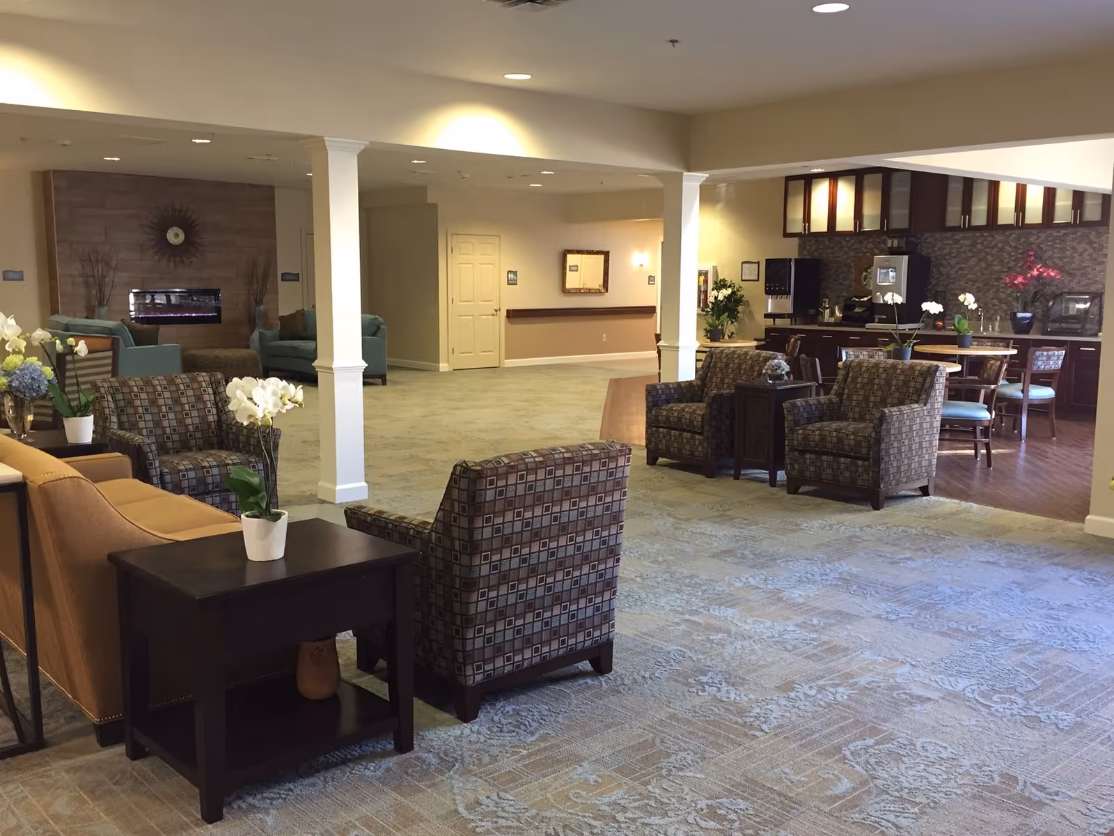 Spacious senior-living common area with armchairs, sofas, side tables, and a kitchenette/dining area in the background.