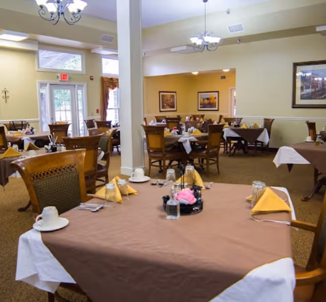 A dining room in an assisted living facility with multiple tables covered in brown and white tablecloths. Each table is set with yellow folded napkins, cups, glasses, and silverware. The room has beige walls, framed artwork, and large windows letting in natural light. Wooden chairs with cushioned seats surround the tables.