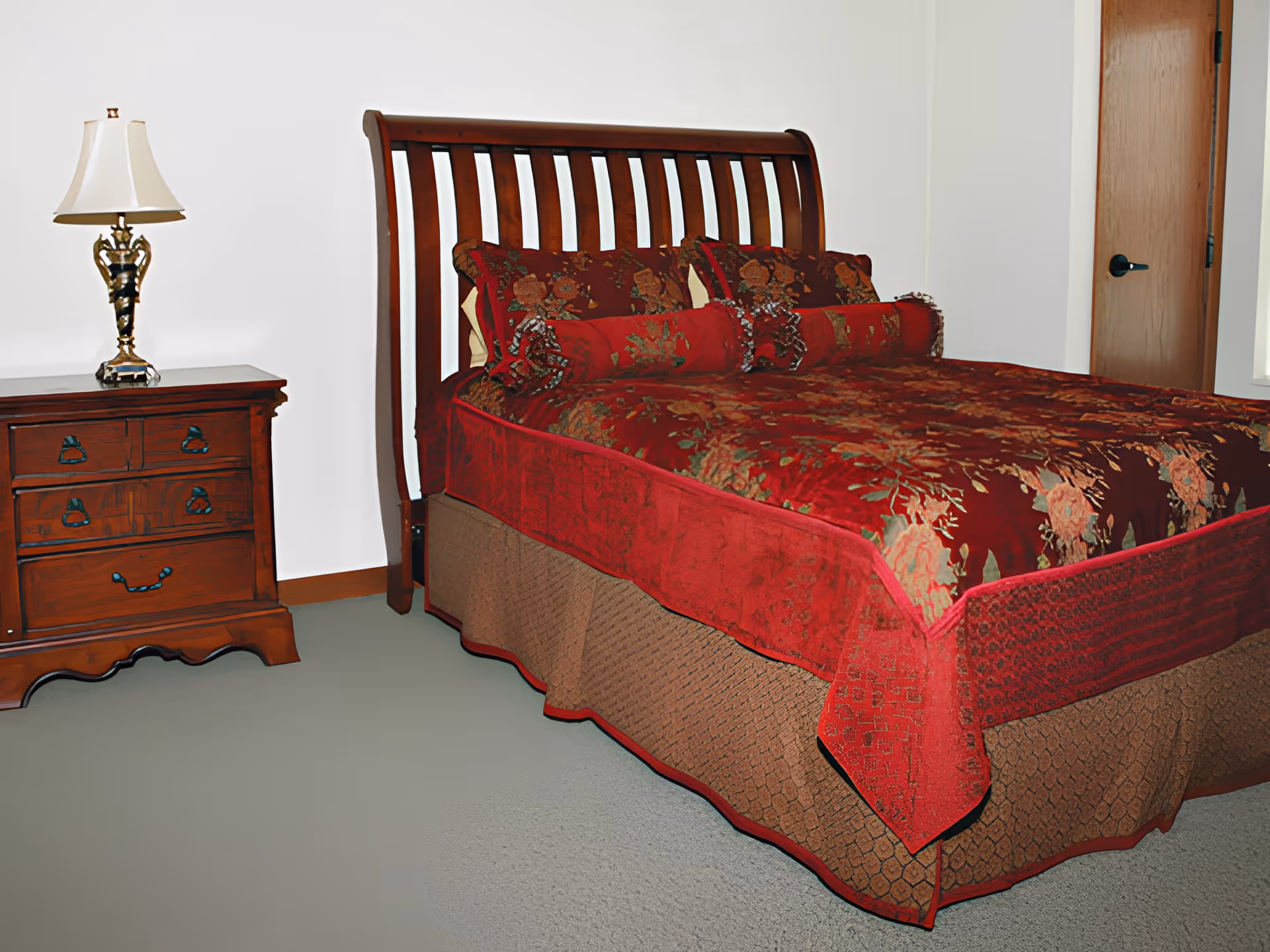 Wooden sleigh bed with a red floral bedspread and matching pillows next to a wooden nightstand with a lamp in a simple bedroom.