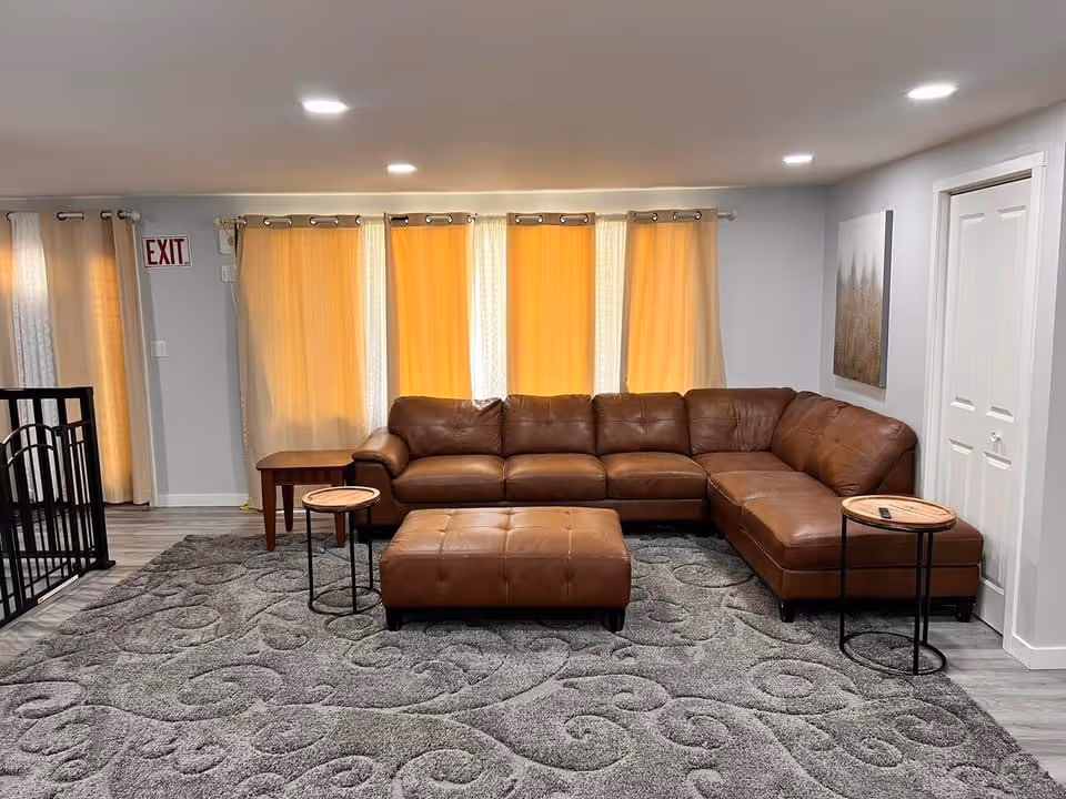 A living room with a large brown leather sectional sofa and matching ottoman on a gray patterned carpet. There are two small round side tables with wooden tops and black metal legs on either side of the sofa. Behind the sofa are three windows covered with beige curtains, and to the left is a door with an exit sign above it. The walls are light gray, and there is a piece of artwork hanging on the right wall. The floor is a light wood laminate.