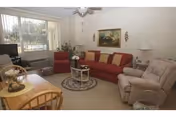 A cozy living room with a red sofa adorned with yellow and red cushions, a beige recliner chair, a small round glass coffee table on a circular rug, a wooden dining table with chairs, a TV on a stand near a window with vertical blinds, a ceiling fan, and framed artwork on the wall.
