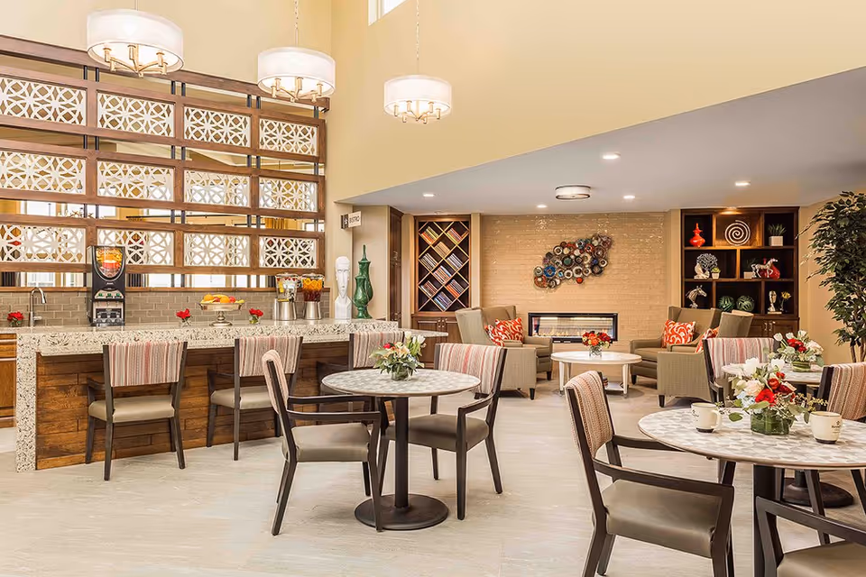 Bright communal dining and lounge area with round tables, a breakfast bar, and a seating area with a fireplace and decorative shelving.