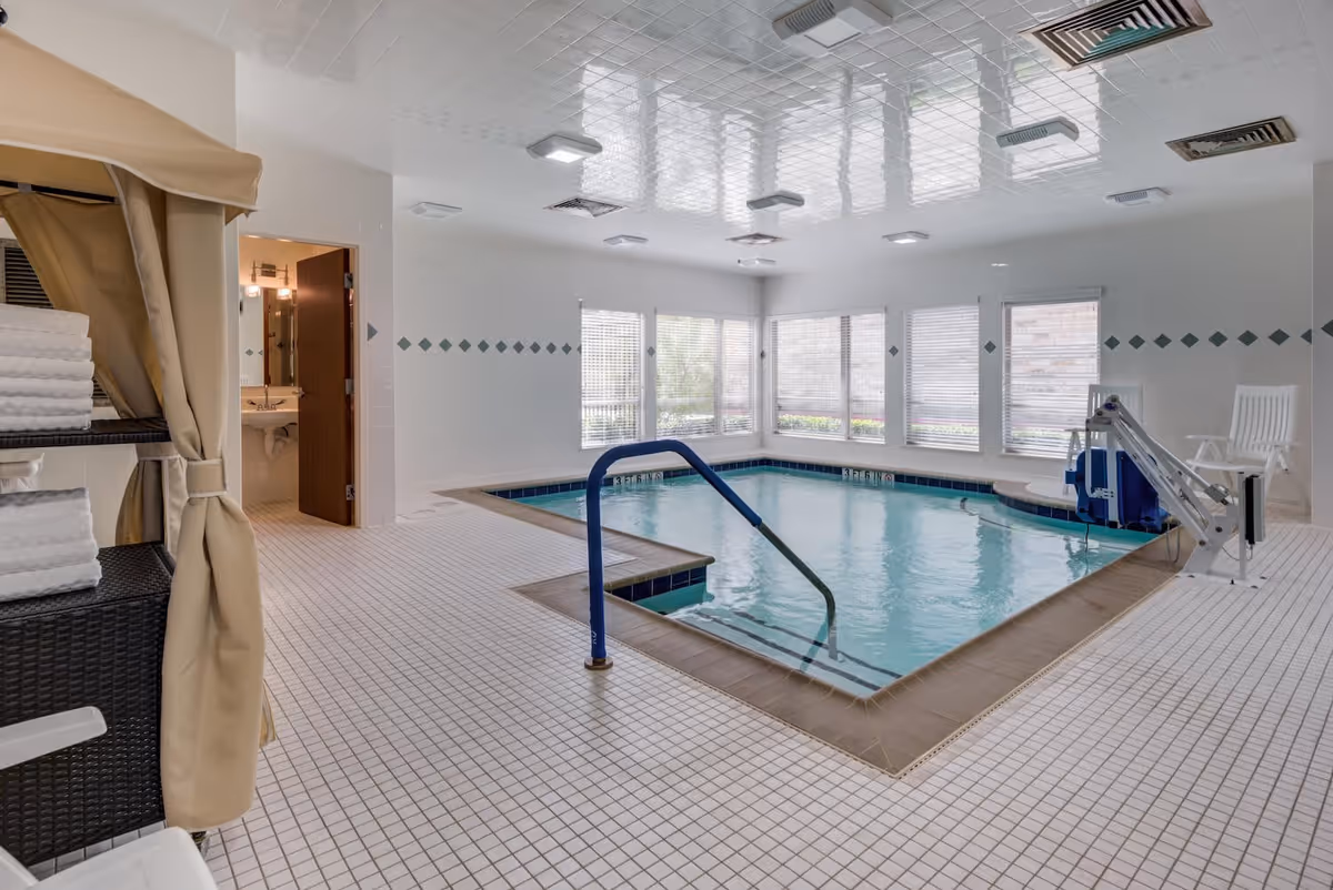 Indoor swimming pool area with tiled floor and walls, a handrail leading into the pool, a pool lift chair for accessibility, and a rack with neatly folded white towels. There is a door leading to a bathroom or changing area, and large windows letting in natural light.