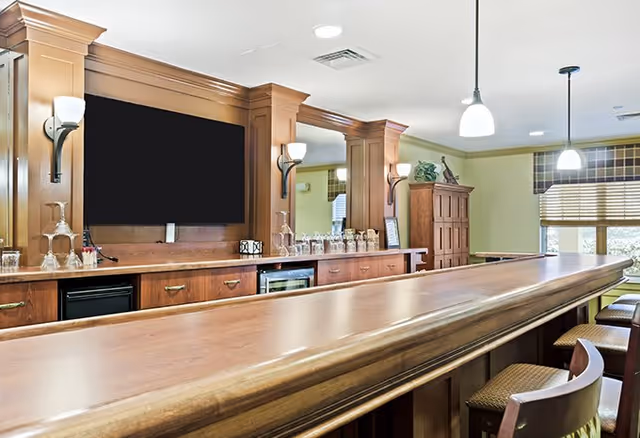 Interior view of a senior living facility's bar area with a long wooden counter, several bar stools, a large flat-screen TV mounted on the wall, and cabinetry with glassware and decorative items. The room is well-lit with pendant lights and wall sconces, and windows with blinds allow natural light in.