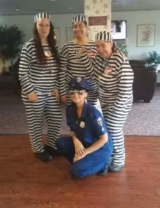 Four people dressed in costumes posing indoors; three are wearing black and white striped prisoner outfits with matching hats, and one is dressed as a police officer in a blue uniform with sunglasses, kneeling in front of the others.