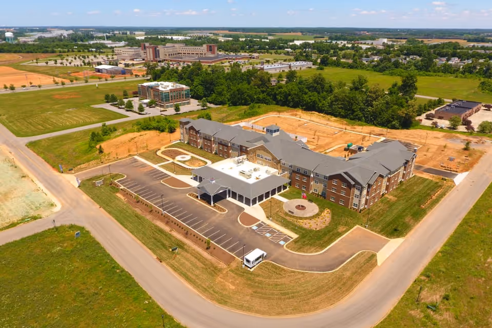 Aerial view of a large senior living facility building with a parking lot, surrounded by green fields and roads. The building has a covered entrance and multiple wings with a gray roof. There are trees and other buildings in the distance under a partly cloudy sky.