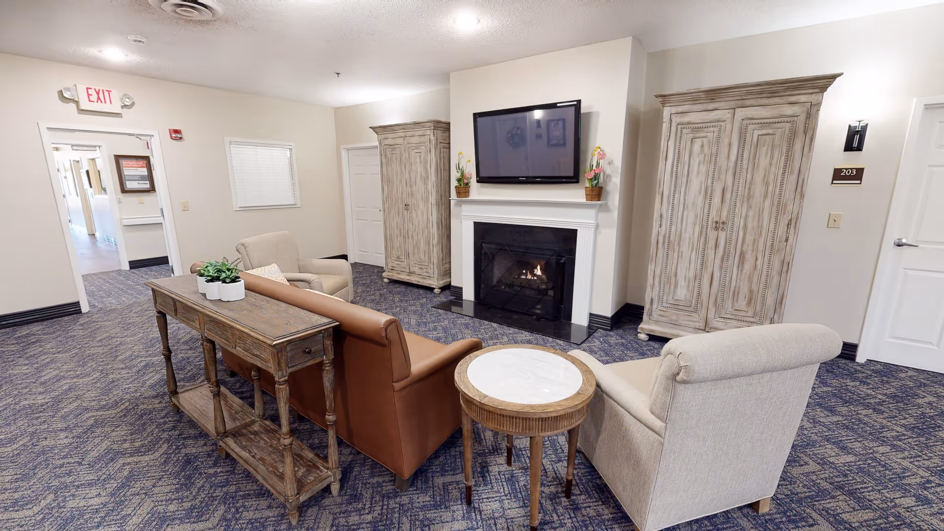 A cozy sitting area in a senior living facility with a brown leather sofa, two beige armchairs, a wooden console table with a plant, a round side table with a marble top, a fireplace with a TV mounted above it, and two large wooden cabinets on either side of the fireplace. The carpet is patterned in blue and beige tones, and there is an exit sign above a doorway leading to a hallway.