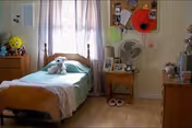 A small bedroom with a single wooden bed covered with a light green blanket and a white teddy bear sitting on it. There is a wooden nightstand next to the bed with a fan, framed photos, and other small items. The room has light-colored walls with some decorations, a window with sheer curtains, and wooden flooring. A pair of slippers is placed on the floor near the nightstand.
