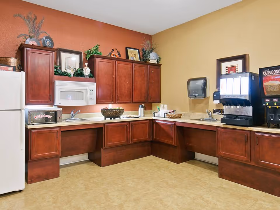 A kitchenette area with wooden cabinets, a white refrigerator, a microwave, a toaster oven, two sinks, a coffee and cappuccino machine, and various decorative items on top of the cabinets and countertop.