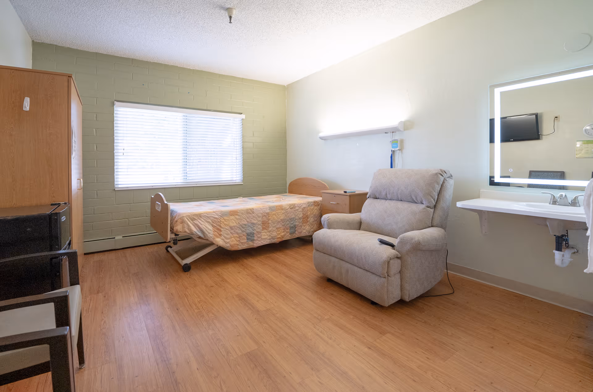 A simple senior care facility bedroom with a single bed covered by a patchwork quilt, a cushioned recliner chair, a wooden cabinet, a small nightstand, a wall-mounted sink with a mirror, and a window with blinds letting in natural light.