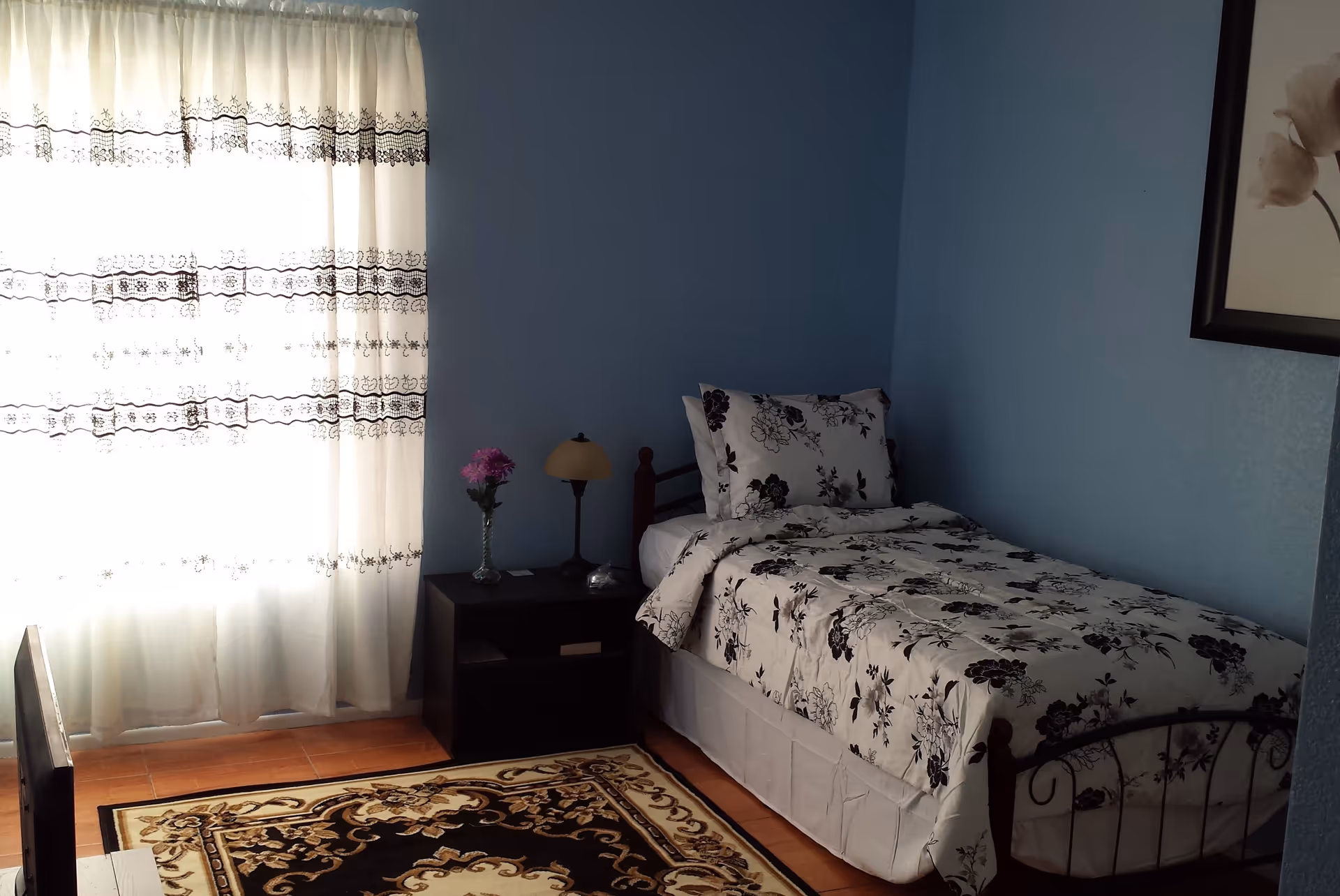 Single-occupancy bedroom with a floral bedspread, nightstand with a lamp and vase, sheer patterned curtains, and a decorative rug.