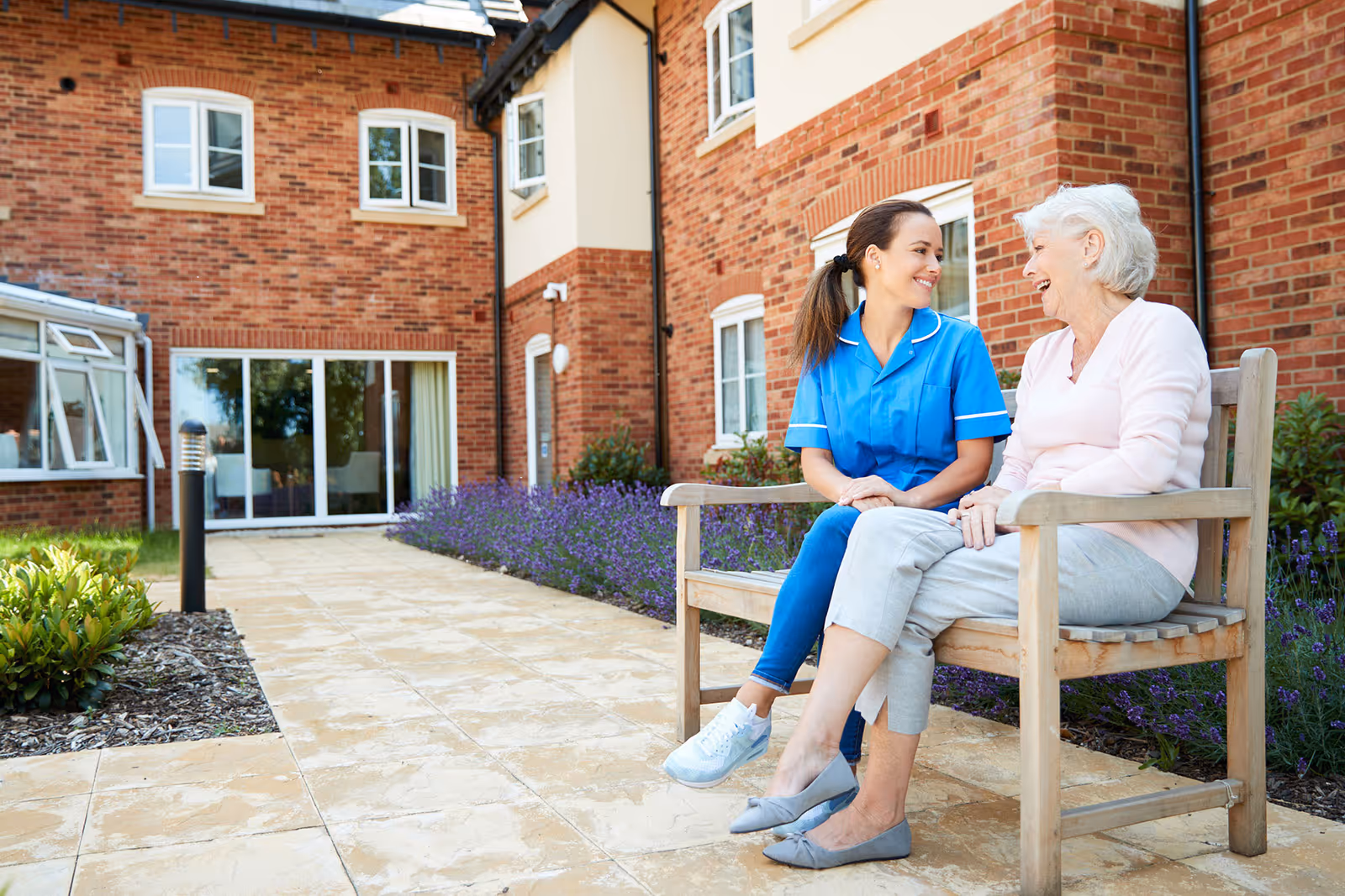 An elderly woman and a caregiver in a blue uniform sitting on a wooden bench outside a brick building, smiling and talking. The area is paved with stone tiles and has flower beds with purple flowers and green shrubs.
