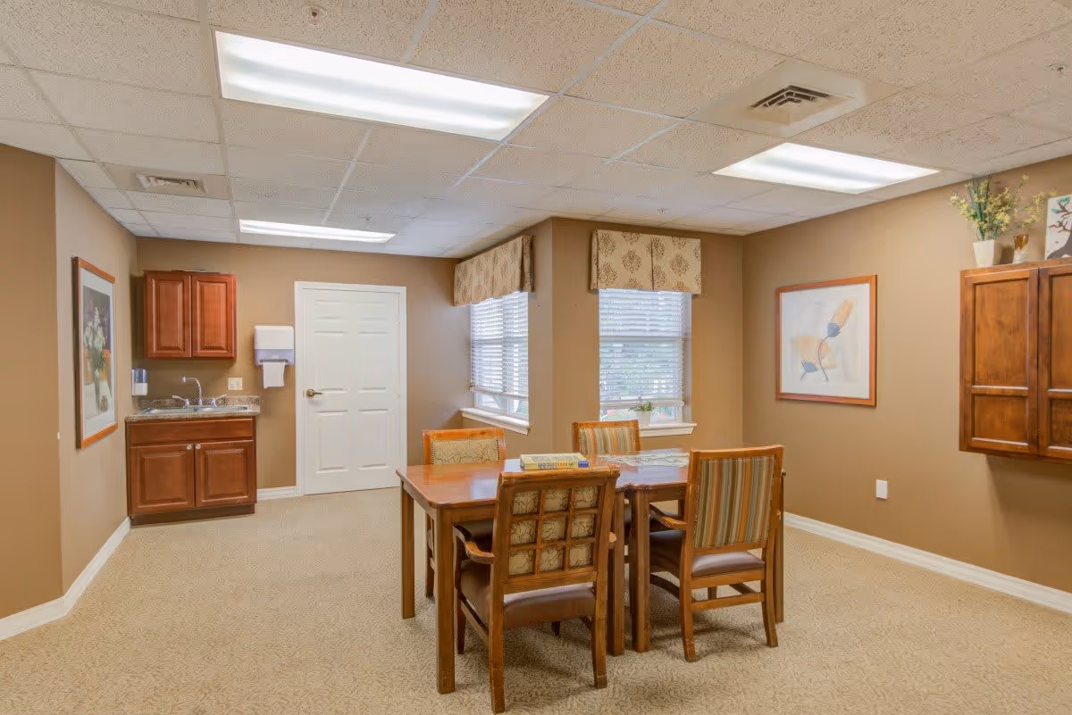 A small dining area in a senior living facility with a wooden table and four chairs. The room has beige walls and carpet, two windows with patterned valances, framed artwork on the walls, and wooden cabinets. There is a small sink and countertop area with a paper towel dispenser on the left side of the room.