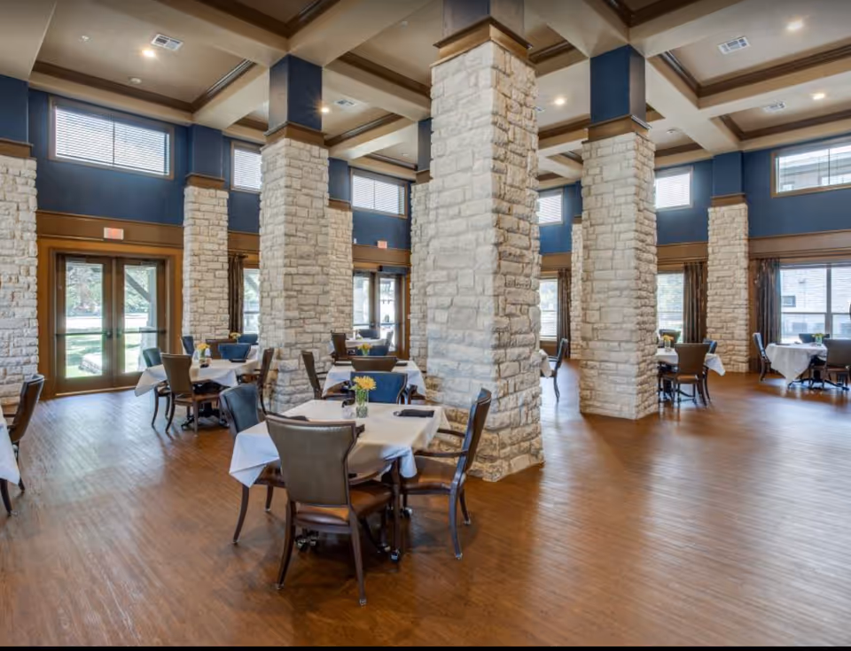 Spacious dining room with multiple tables covered with white tablecloths and set with chairs. The room features large stone pillars, high ceilings with exposed beams, and large windows letting in natural light. The floor is wooden, and there are small flower vases on each table.