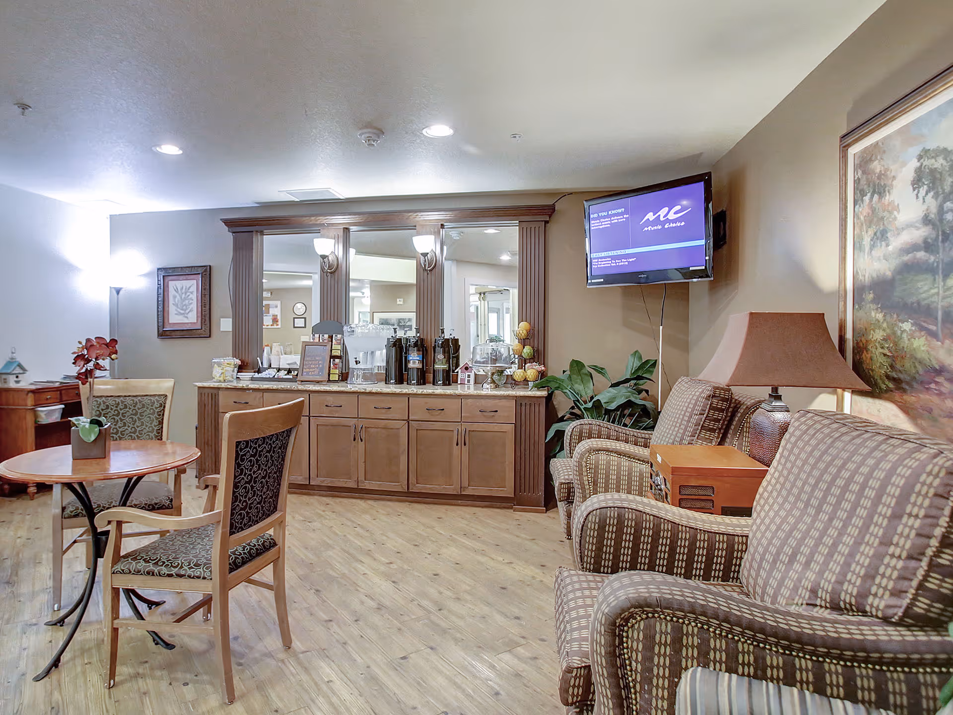 Seating area in a senior living common room with armchairs, a small table, and a coffee station under a mirrored cabinet.