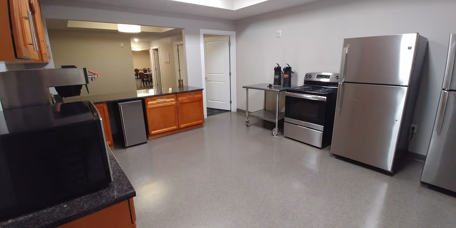 A spacious kitchen area with stainless steel appliances including a refrigerator and stove. There are wooden cabinets with a dark countertop, a microwave, and a small refrigerator under the counter. Two coffee dispensers are placed on a metal rolling cart next to the stove. The kitchen opens into a hallway leading to a dining area with tables and chairs.