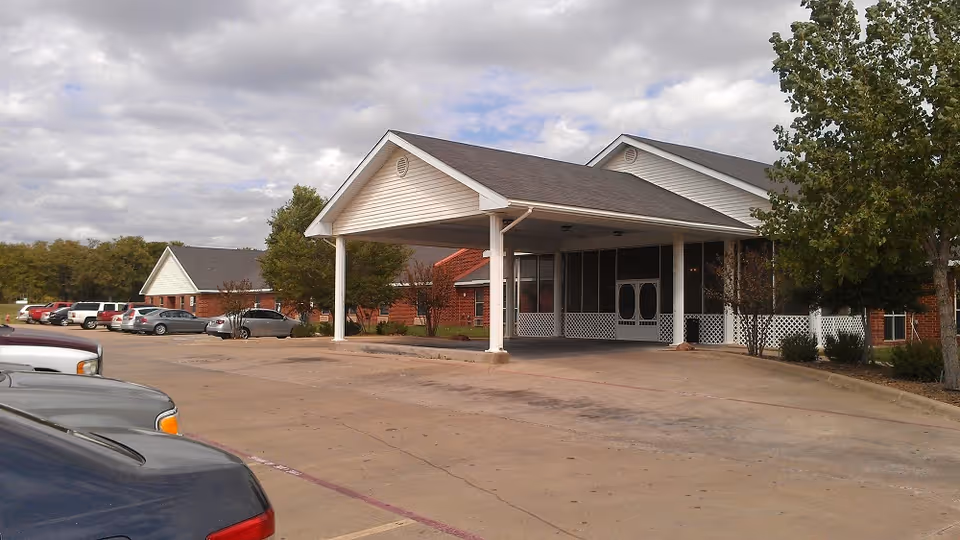 Front entrance of a single-story brick nursing and rehabilitation facility with a covered porte-cochere, parking lot and several parked cars under a cloudy sky.