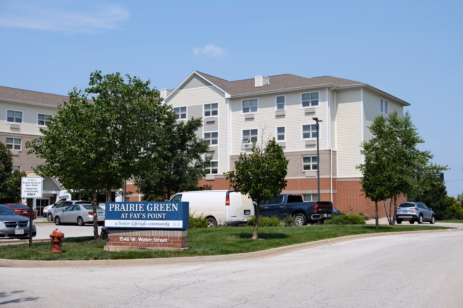 Exterior view of the Prairie Green at Fay's Point senior living building with a front sign, trees, and parked cars.