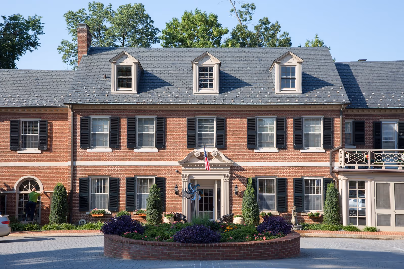 Brick colonial-style building front with a central entrance, dormer windows, and a landscaped circular planter.