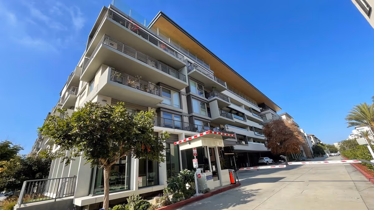 Modern multi-story senior living building with balconies, a gated driveway and entrance booth under a clear blue sky.