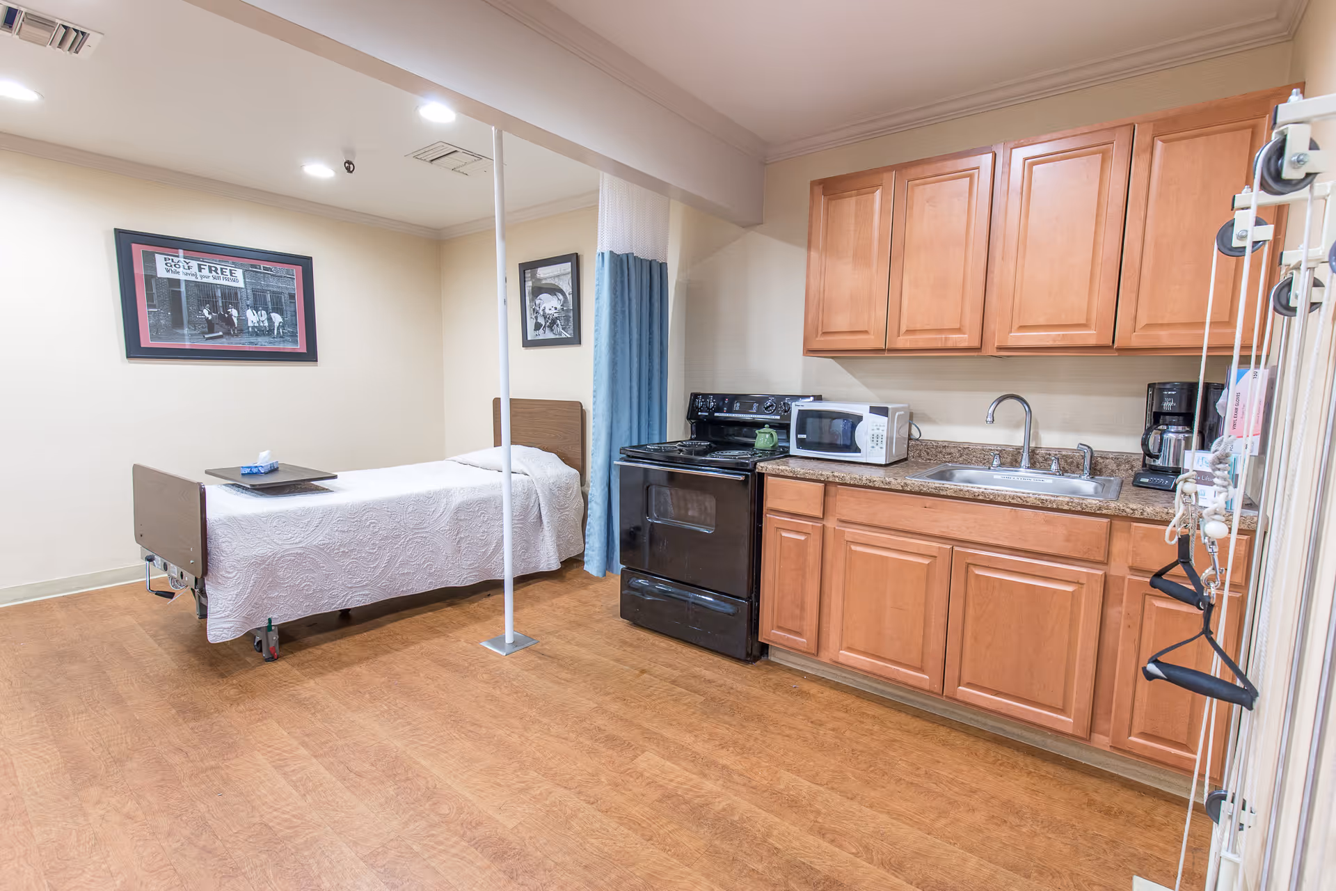 A room with a single hospital-style bed covered with a white quilt, a kitchenette with wooden cabinets, a black stove, microwave, coffee maker, and a sink. The room has light-colored walls, wood flooring, and two framed pictures on the walls. A blue privacy curtain is partially visible near the bed.