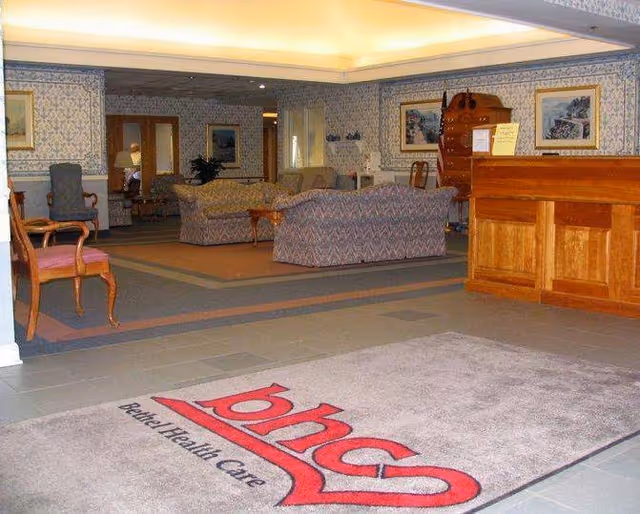 Interior view of a senior living facility lobby with a large carpet displaying the Bethel Health Care logo. The room features a wooden reception desk on the right, patterned sofas and chairs arranged in a seating area, framed artwork on the walls, and soft overhead lighting.
