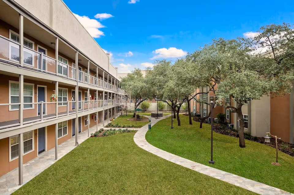 Outdoor courtyard area of a senior living facility with a curved stone pathway, green grass, several trees, and a two-story building with balconies and doors overlooking the courtyard under a blue sky with some clouds.
