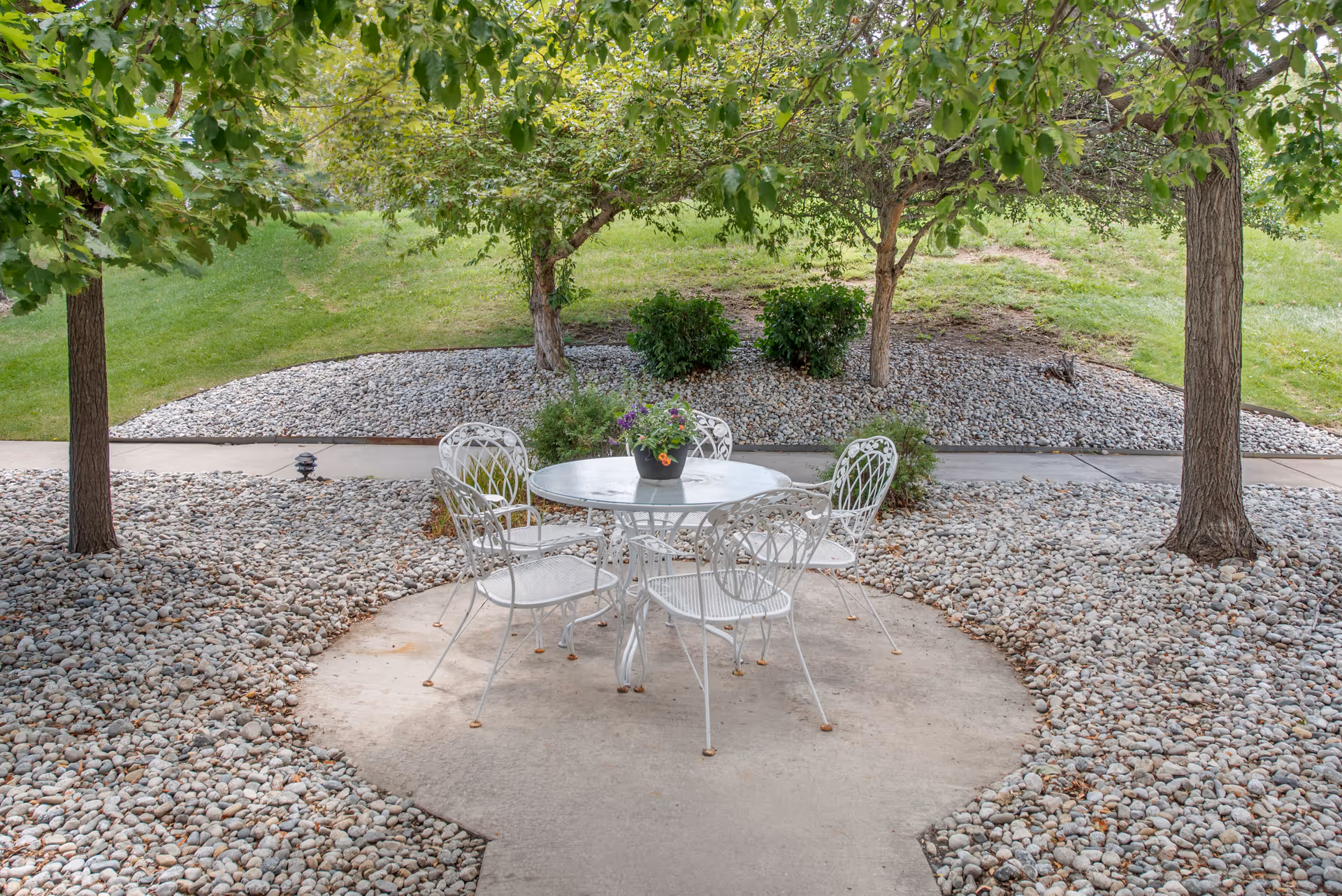Outdoor patio with a white metal table and four chairs under trees surrounded by decorative rocks and a walkway.