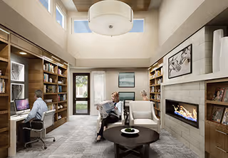 A cozy senior living common area with a man working on a computer at a built-in wooden desk on the left, and a woman sitting comfortably in a chair reading a newspaper near a modern fireplace. The room features tall ceilings with a large circular light fixture, built-in bookshelves, a round coffee table, and large windows letting in natural light.