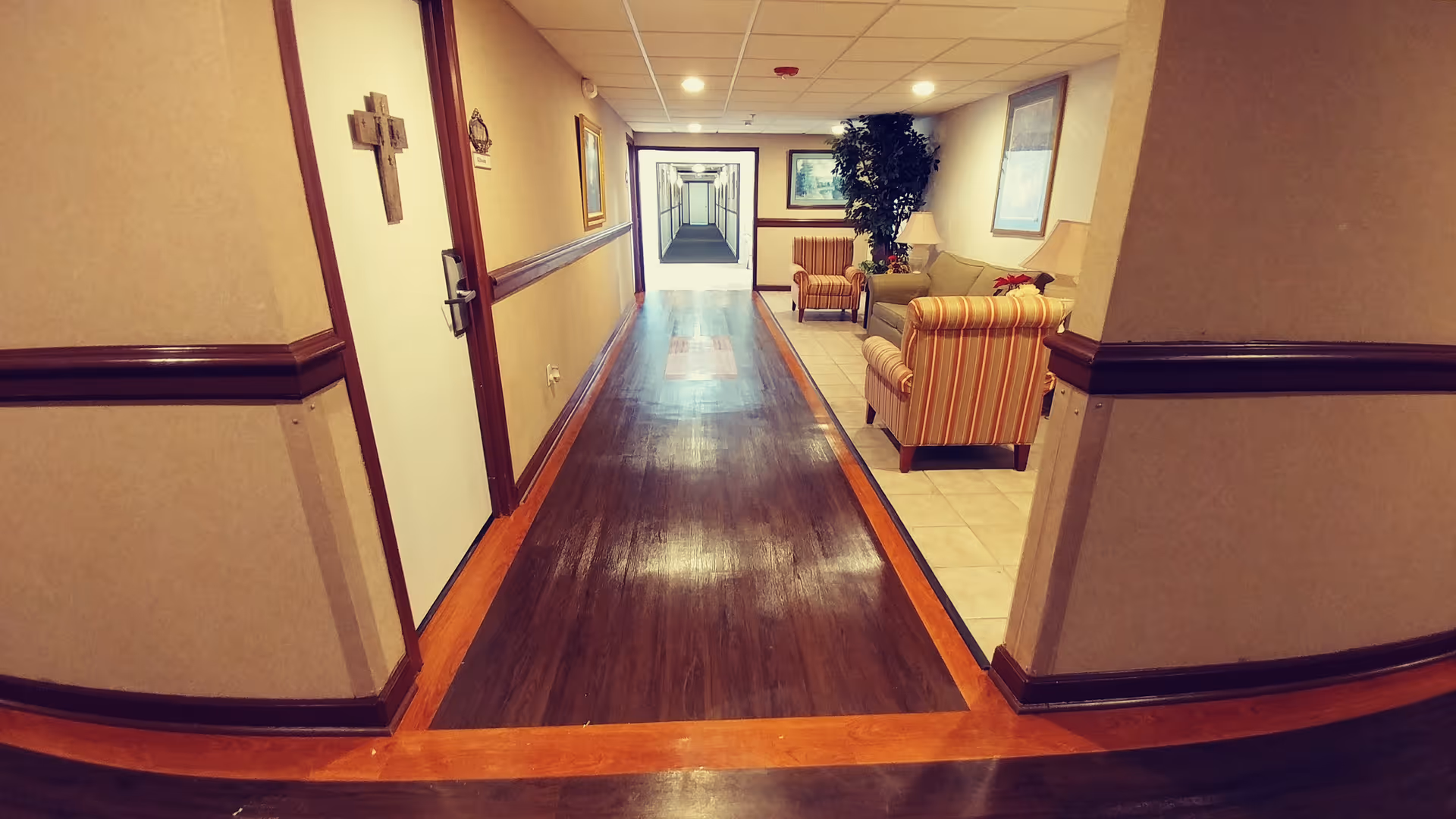 A long hallway in a senior living facility with polished wooden floors and beige walls. On the right side, there is a small seating area with striped armchairs, a sofa, a lamp, and a potted plant. The hallway extends into the distance with doors and framed pictures on the walls.