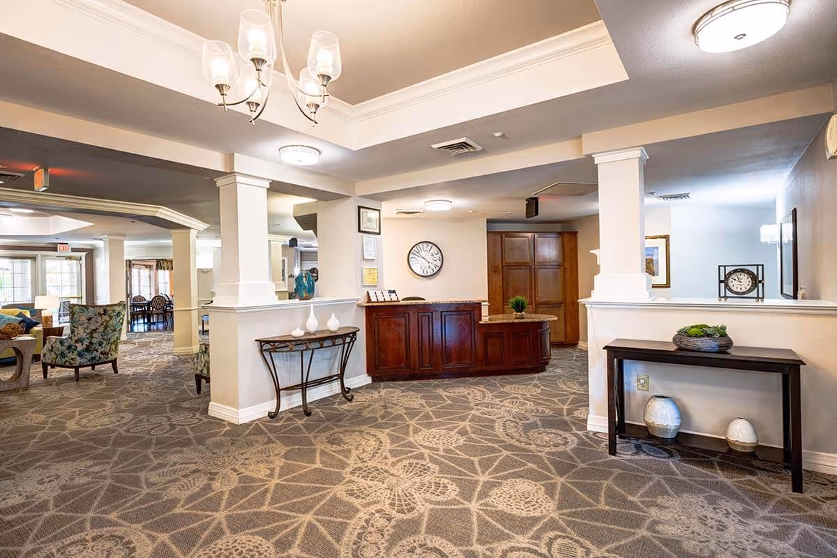 Interior view of a senior living facility lobby area with a wooden reception desk, decorative tables with vases, patterned carpet, and seating area with armchairs in the background.