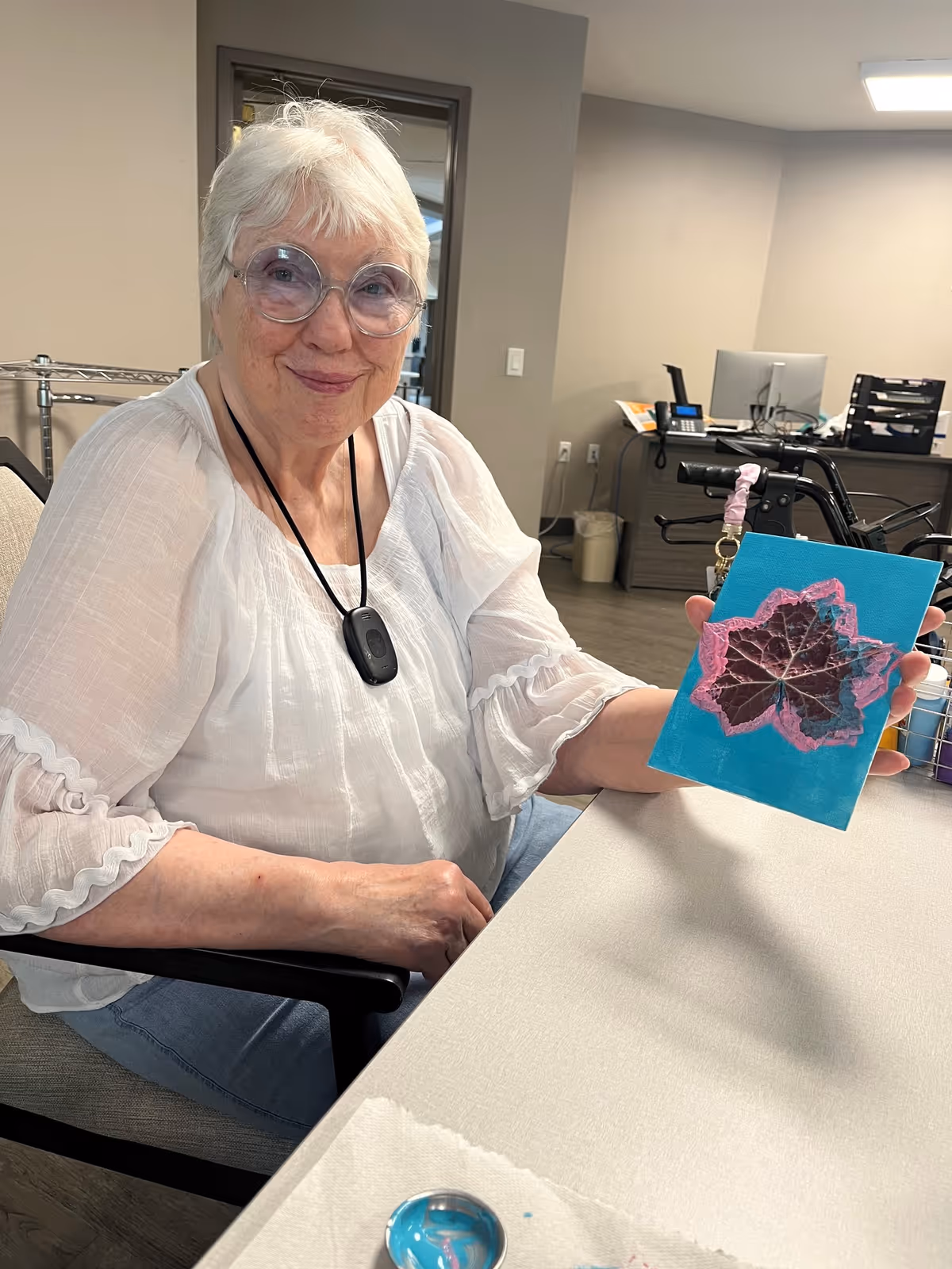 An elderly woman with short white hair and glasses is sitting at a table in a room. She is wearing a white blouse and a black medical alert necklace. She is holding up a small blue canvas with a painted leaf design in pink and brown. In the background, there is office furniture including a desk, computer, and a walker.