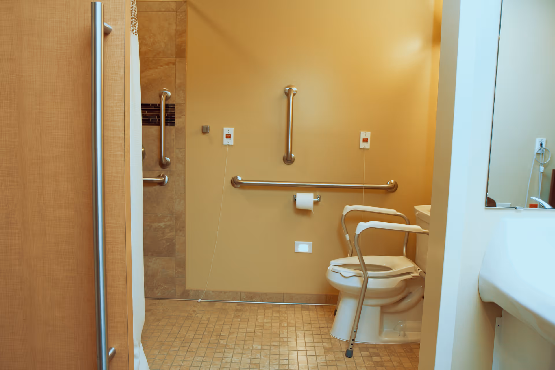 Accessible bathroom with a toilet equipped with safety rails, grab bars on the walls, emergency pull cords, and a tiled floor. A sink and mirror are partially visible on the right side.