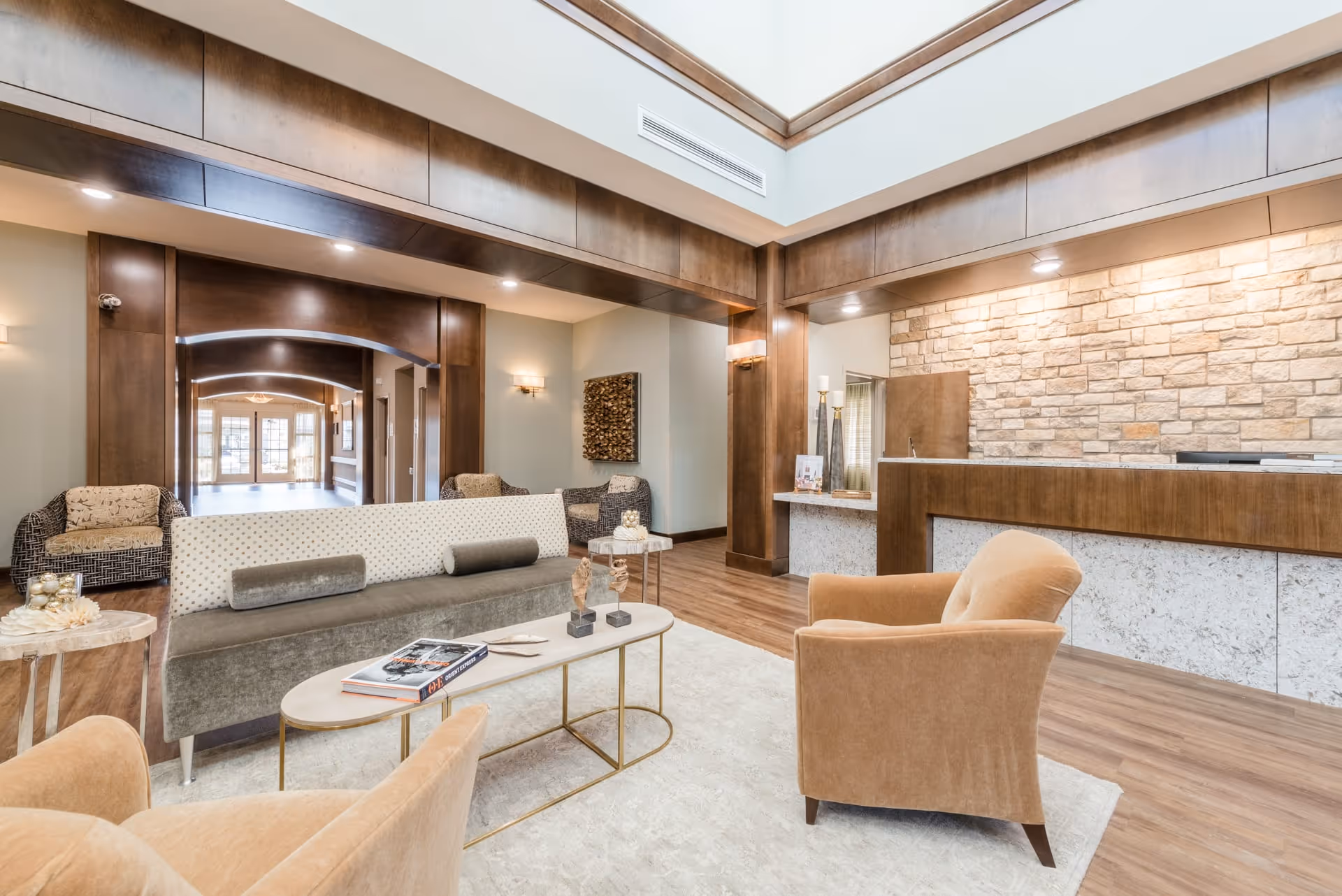 A bright and spacious nursing facility lobby with comfortable seating including armchairs and a sofa around a coffee table. The reception desk is visible on the right with a stone wall backdrop. The area features wooden accents, recessed lighting, and a high ceiling with a skylight.