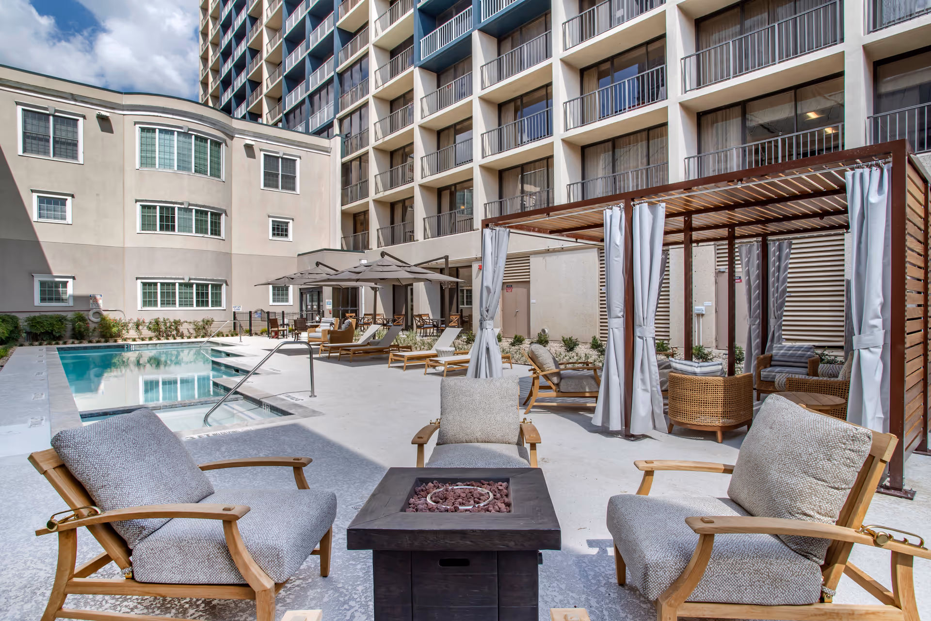 Outdoor courtyard with a swimming pool, lounge chairs, cabanas, and cushioned seating with a fire pit in front of a multi-story building with balconies.