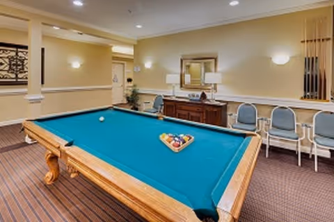 A well-lit indoor recreational room featuring a pool table with a teal felt surface, a set of billiard balls arranged in a triangle, and a cue ball. The room has beige walls with wall-mounted lights, a wooden sideboard with two table lamps and a mirror above it, and several blue cushioned chairs lined up against the wall. There is a rack with pool cues on the right side of the image.