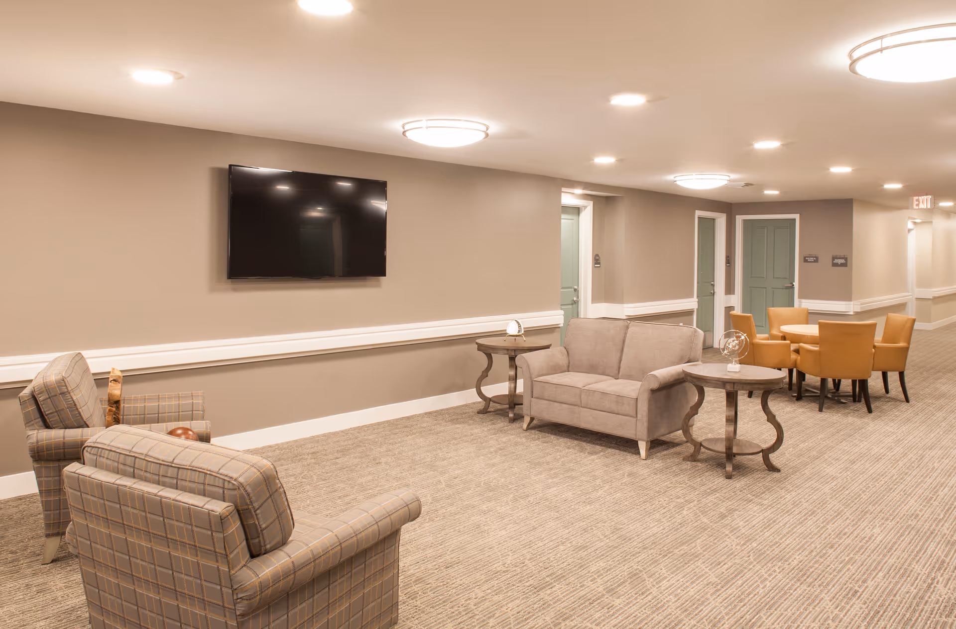 A well-lit common area in a senior living facility featuring a wall-mounted flat screen TV, two plaid armchairs, a beige loveseat, two round side tables, and a round table with four tan leather chairs. The walls are painted beige with white trim, and there are several doors along the hallway.